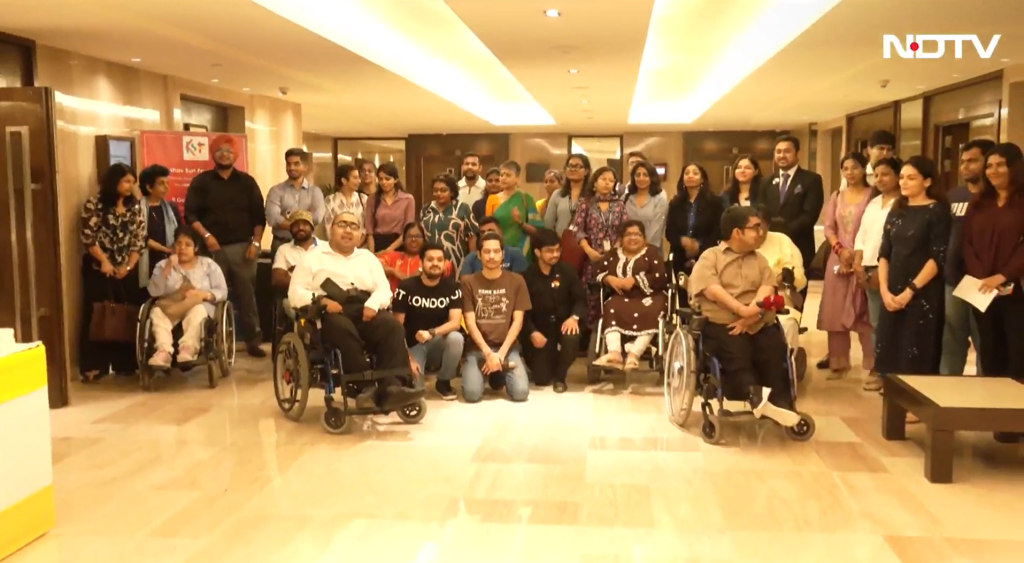 A large group of people, including several individuals in wheelchairs, poses for a photograph in a brightly lit indoor corridor