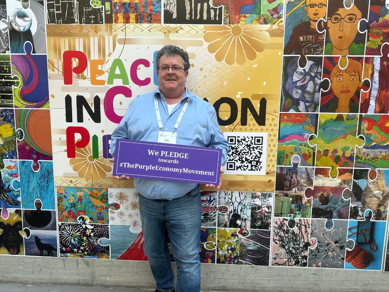 Person holding a Purple Economy pledge sign in front of a colorful puzzle mural.