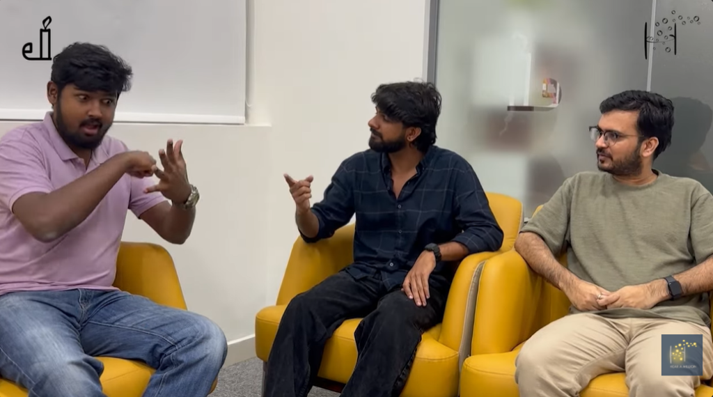 Three people sit on yellow chairs, with one using sign language while the other two engage in the conversation.