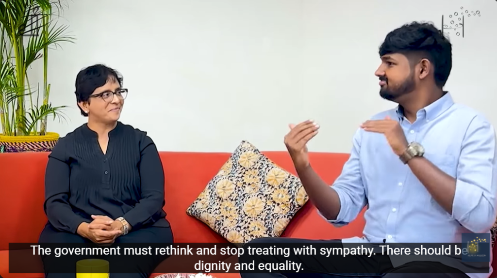 A presenter uses sign language while speaking with Shanti Raghavan on a red sofa.