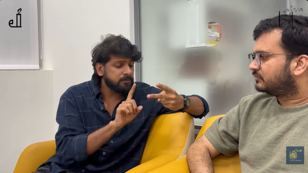 Two people sit on yellow chairs, with one using sign language while the other listens attentively.